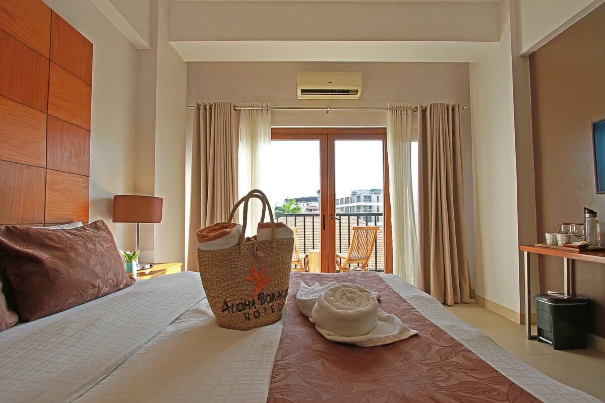 A hotel room with a bed, a window with curtains, a balcony view, a trash bin, and a bag with the hotel name.