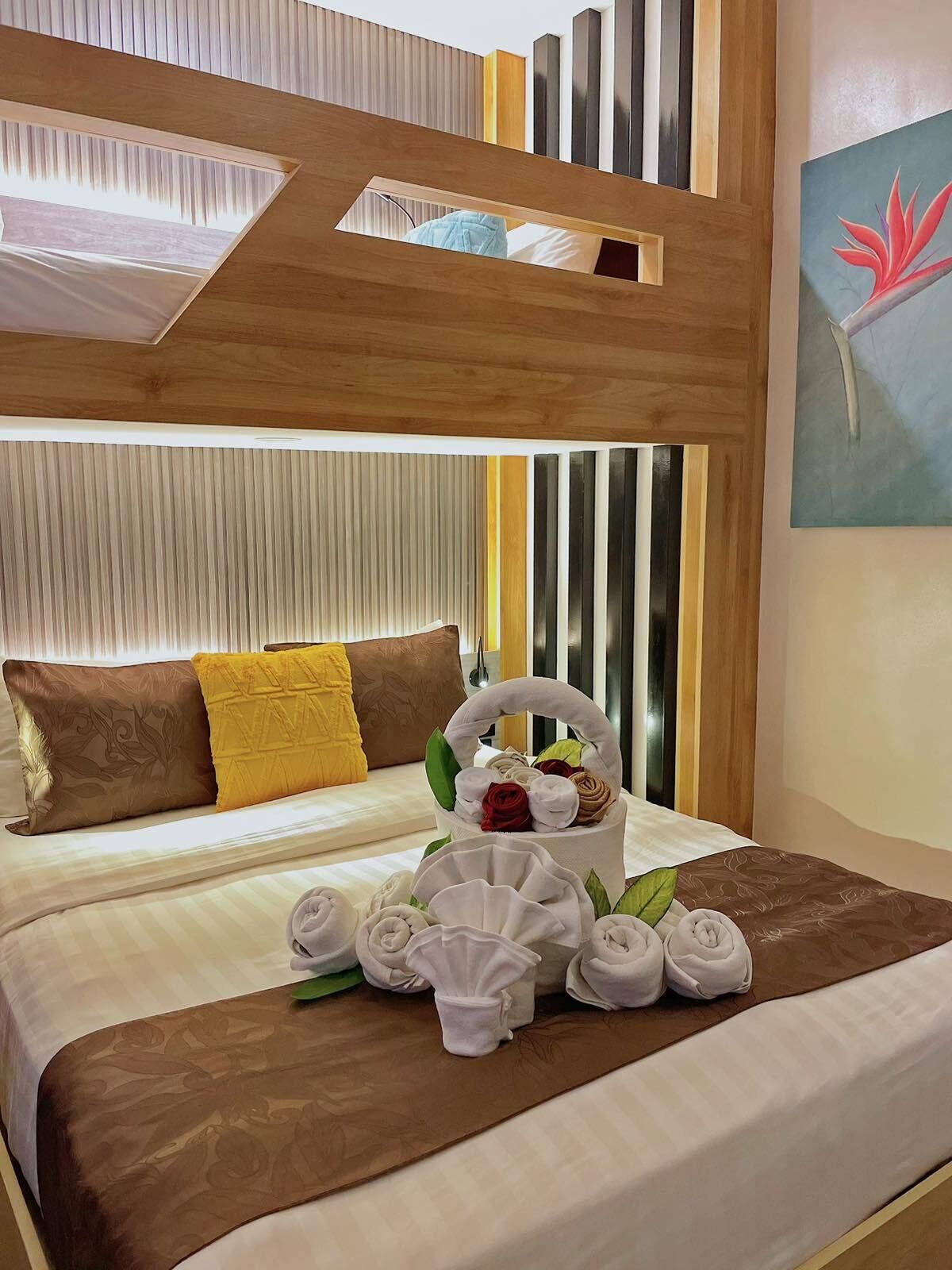 A hotel room with a bunk bed, a bedside table with towels and a basket of toiletries, and a painting on the wall.