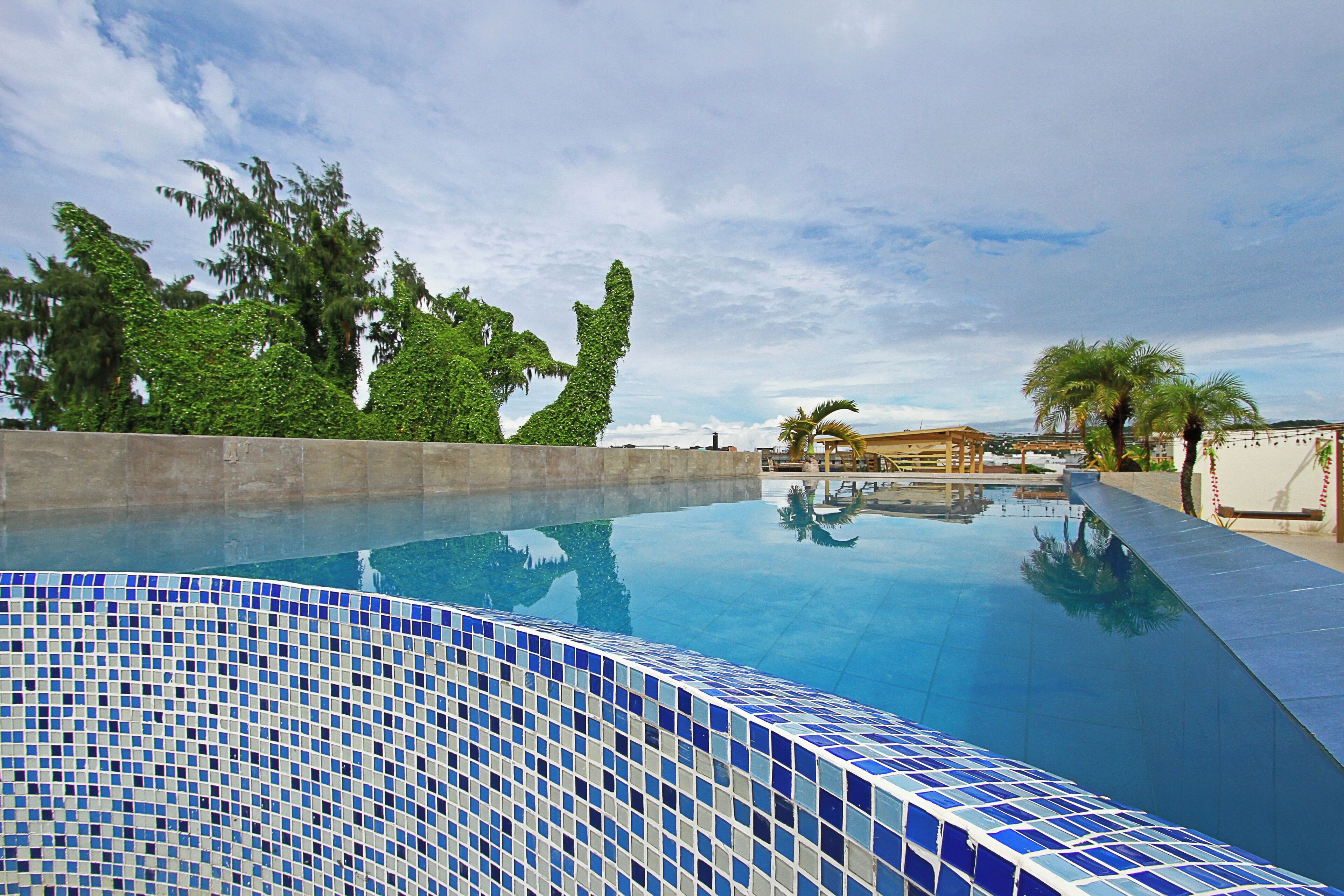 A swimming pool with blue mosaic tiles, surrounded by greenery and a clear sky.