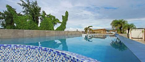 Una piscina con mosaicos azules, rodeada de follaje y un cielo despejado.