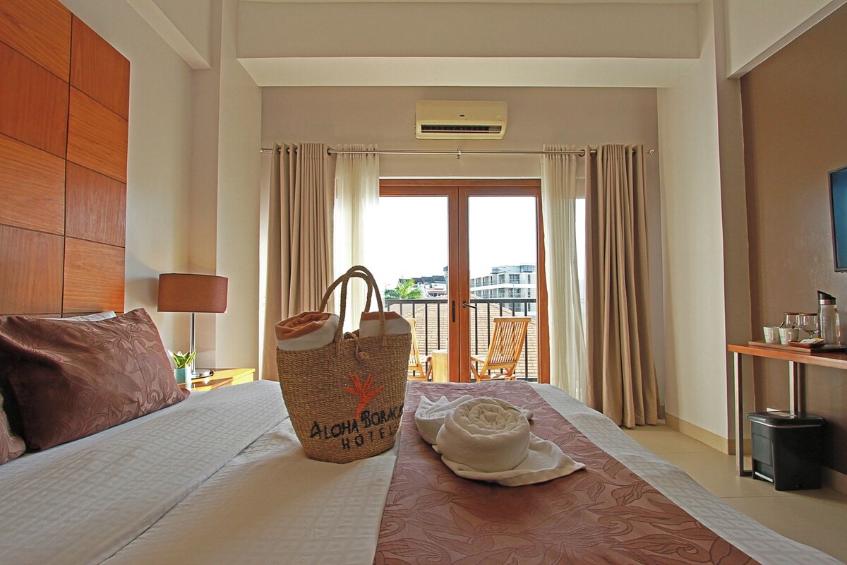 Une chambre d’hôtel avec un lit, une grande fenêtre, des rideaux, un appareil de climatisation, une chaise en bois et une table avec une bouteille et des verres.