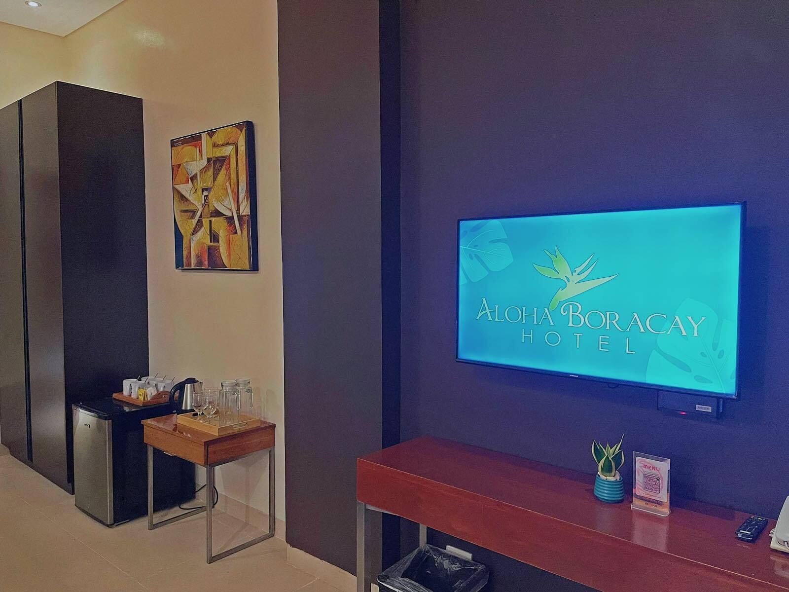 Zona de recepción con un televisor mostrando el logotipo de Aloha Boracay Hotel, una mesa pequeña con una planta y un cubo de basura.