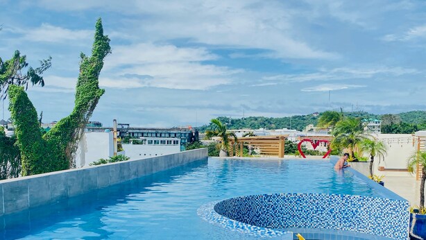 Una piscina con borde de azulejos, rodeada de vegetación y con vistas a edificios al fondo.