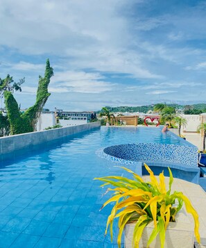 Una piscina con borde de azulejos, rodeada de vegetación y con vistas a edificios al fondo.