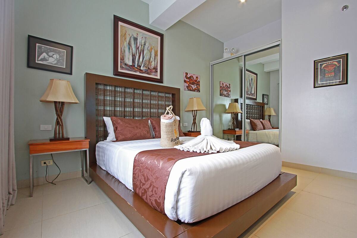 A modern hotel room with a large bed, a wooden headboard, a bedside table with a lamp, and a large mirror.