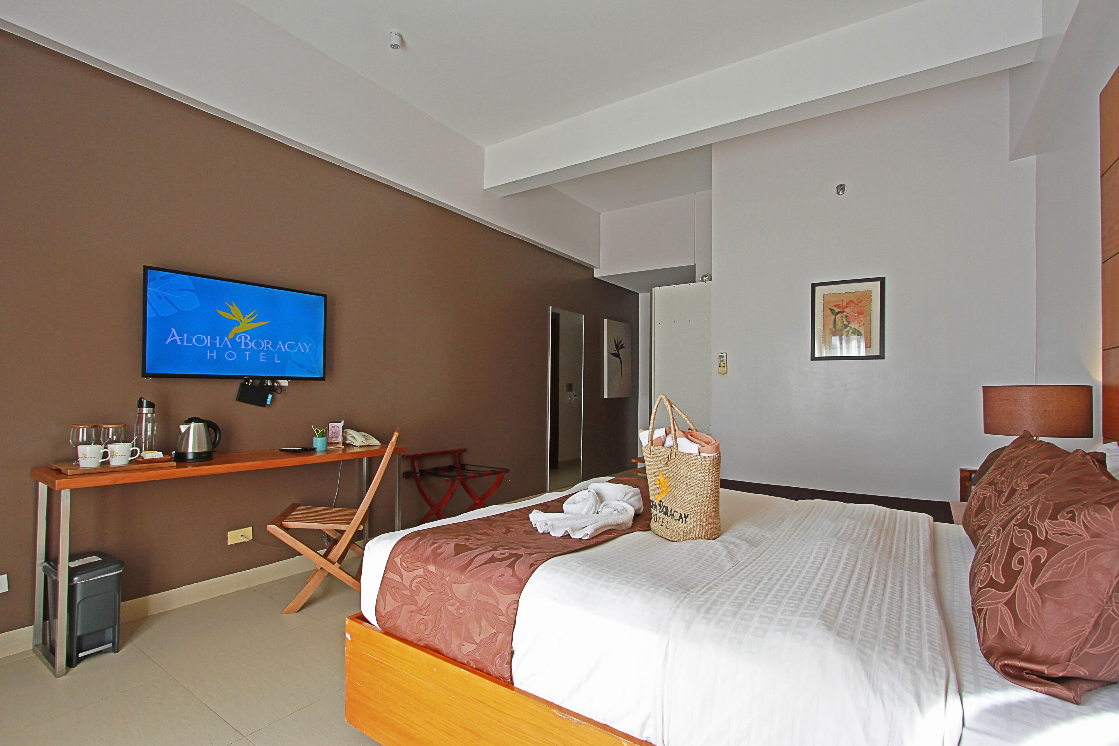 A hotel room with a bed, a desk with a television displaying the hotel name, a trash bin, and a coat rack.