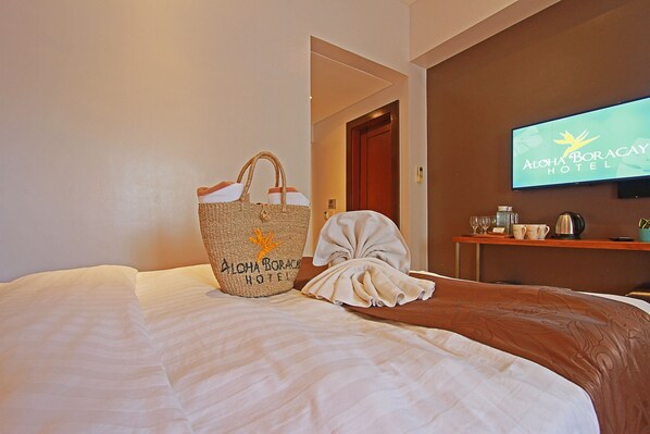 Habitación de hotel con una cama, un bolso tejido grande, una bata y un televisor mostrando el nombre del hotel.