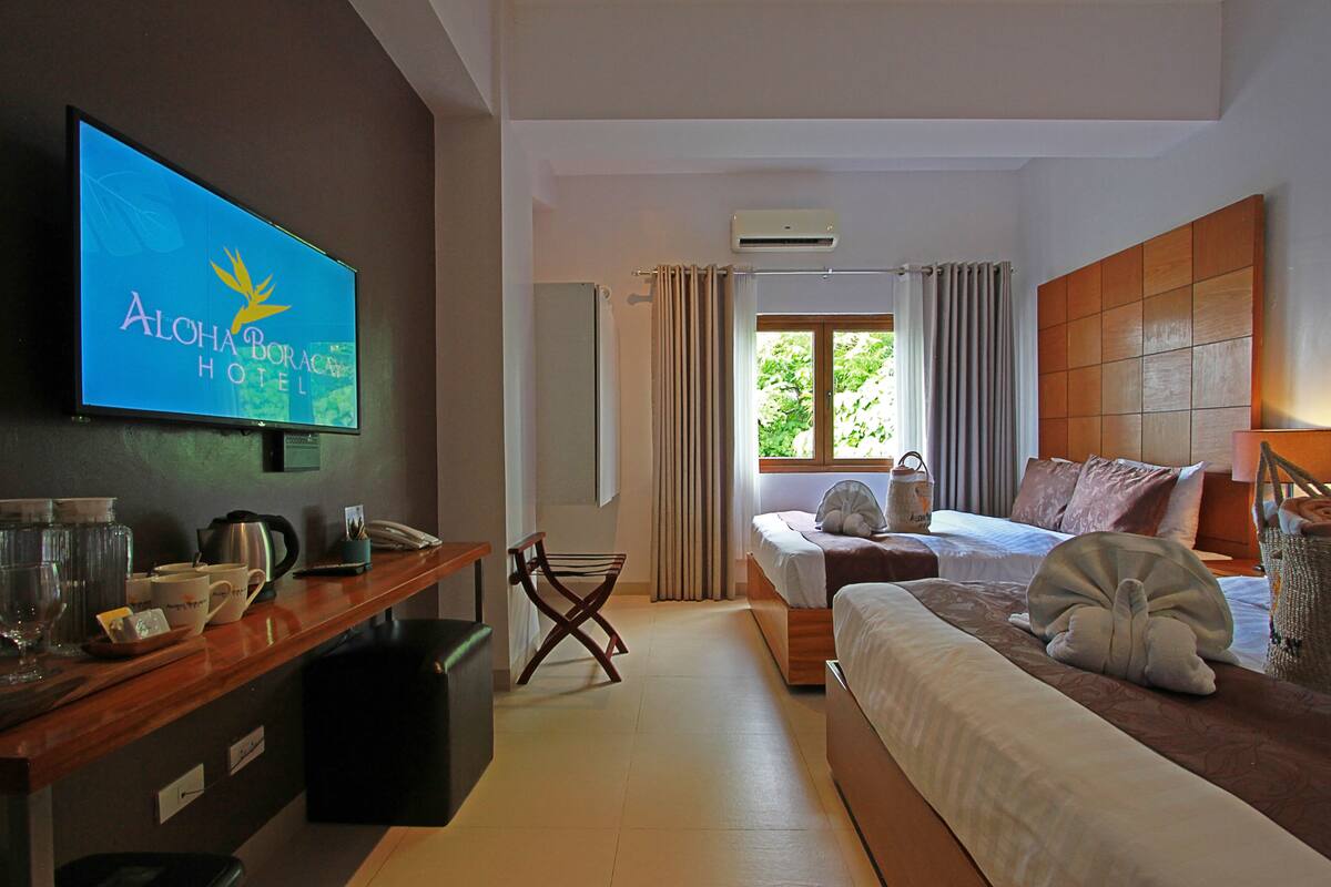 A hotel room with a bed, a TV mounted on the wall, a window with curtains, and a wooden desk with a kettle and cups.