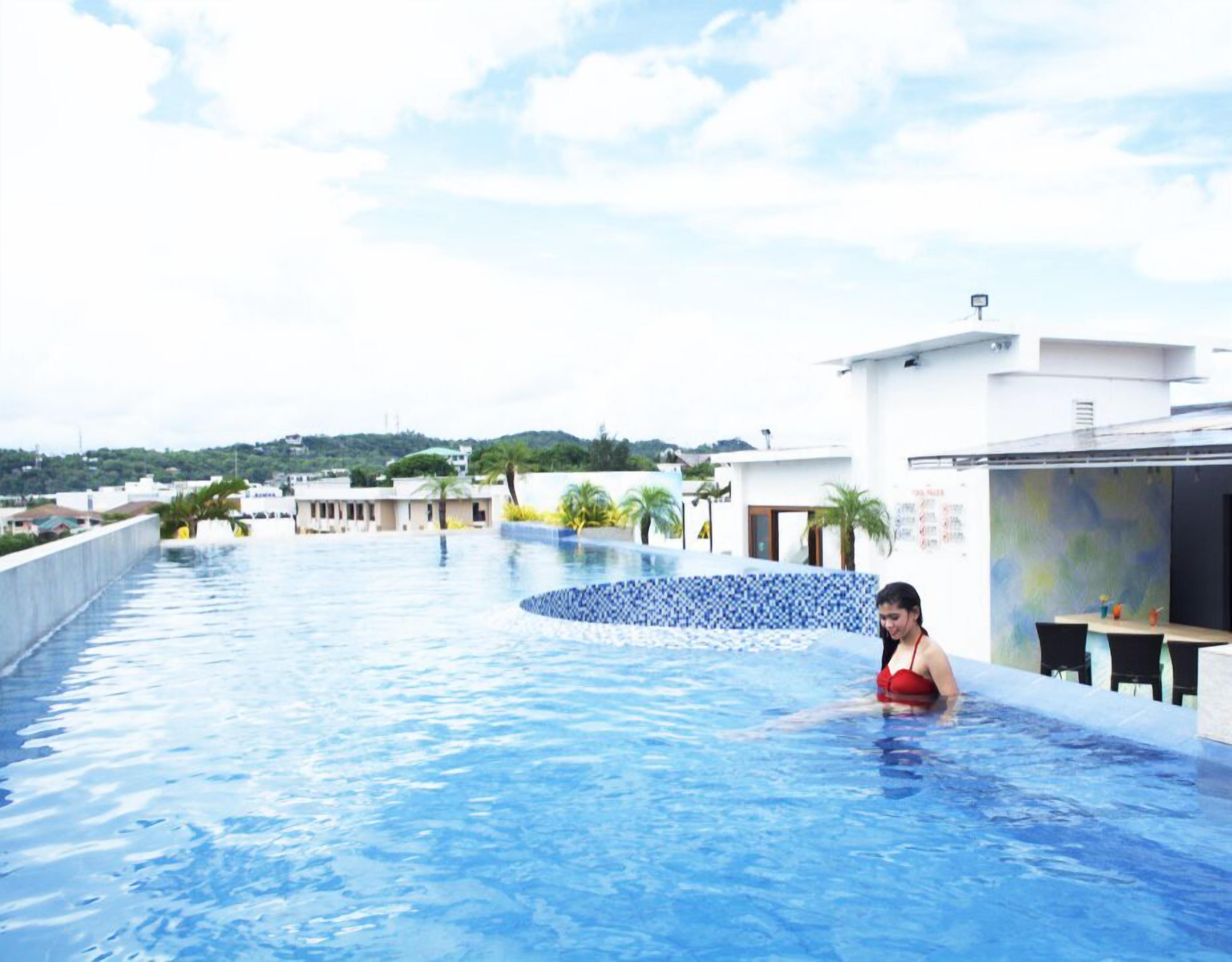Ein Pool mit einer gefliesten Kante, eine Person im roten Badeanzug und im Hintergrund ein Blick auf Gebäude und Grünflächen.