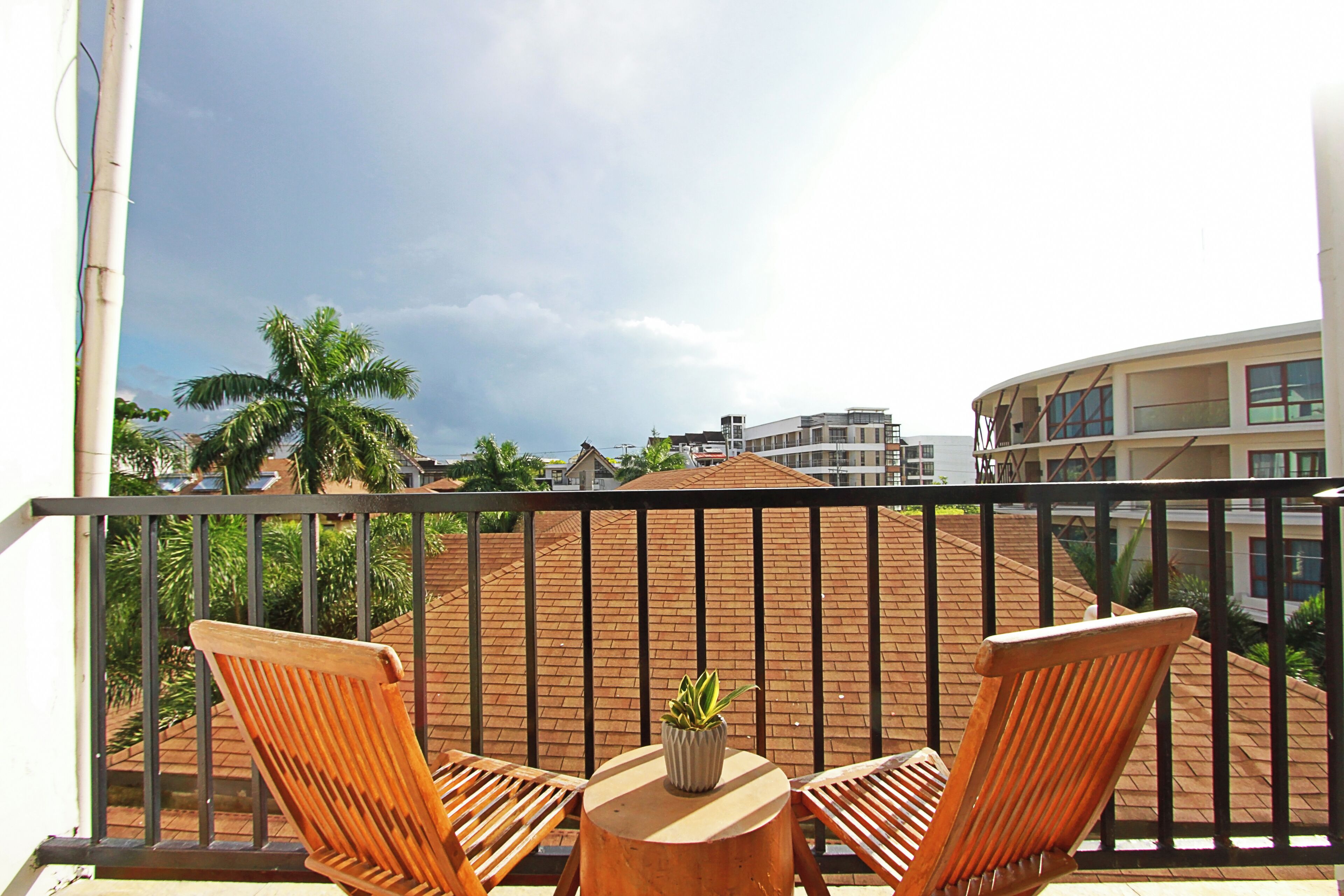 A balcony with two wooden chairs and a small round table, a potted plant, and a view of a building with a red roof and palm trees.