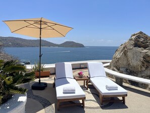 A seaside terrace with two sun loungers, a large umbrella, and a view of the sea and hills. - Villa de la Roca (Zihuatanejo)