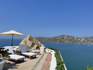 Un área de descanso junto al mar con sillones y una sombrilla, con vistas a un mar tranquilo y un pueblo costero.