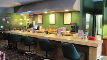 Un comptoir avec une machine à café, des panneaux affichant le menu et un panneau indiquant la zone où passer les commandes. On peut voir des tabourets de bar et une poubelle au premier plan.