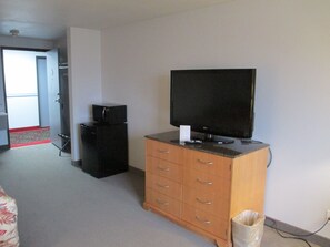 Quarto com televisão sobre um móvel de madeira, um frigorífico preto pequeno e uma porta que dá para outro espaço.