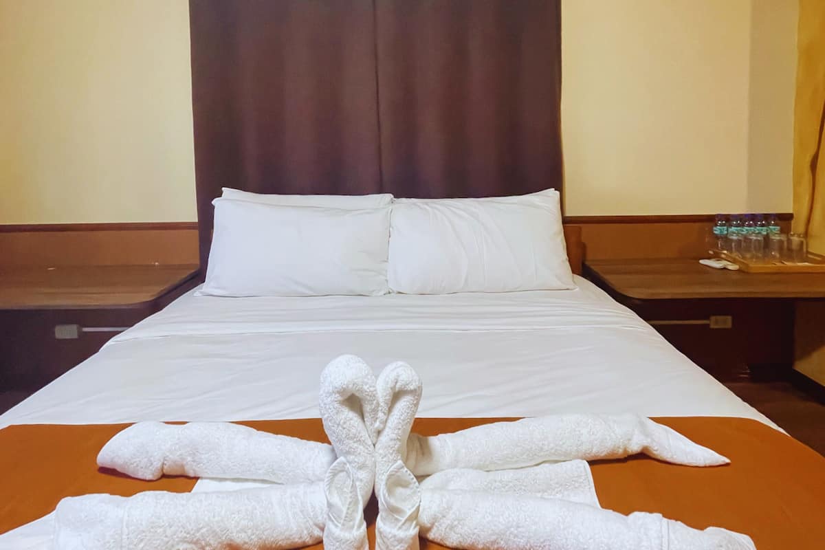 A hotel room with a large bed, two pillows, a wooden nightstand with a tray of water bottles, and towels arranged in a heart shape on the bed.
