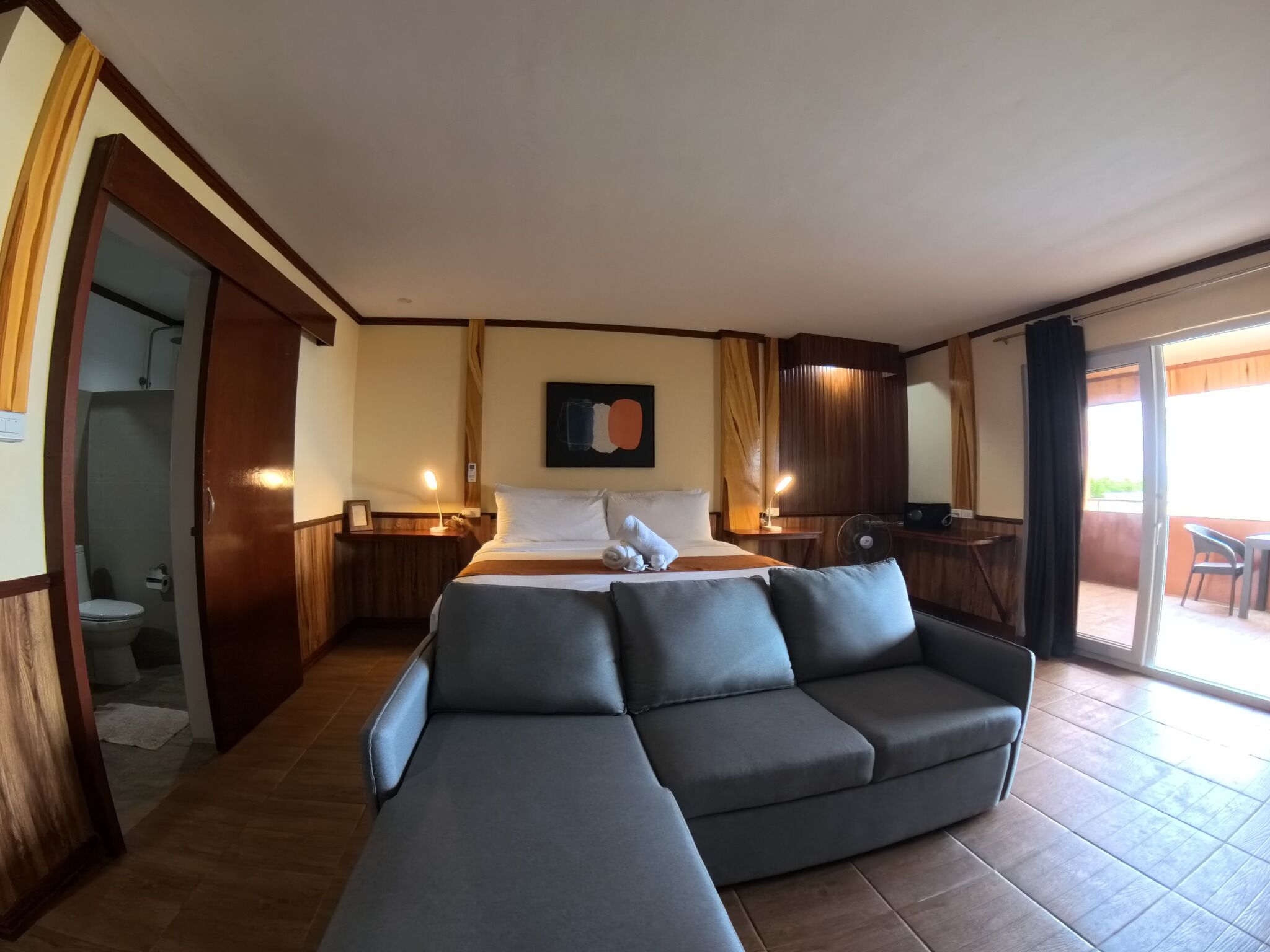 A hotel room with a bed, a sofa, a small table, a fan, and a sliding glass door leading to a balcony.