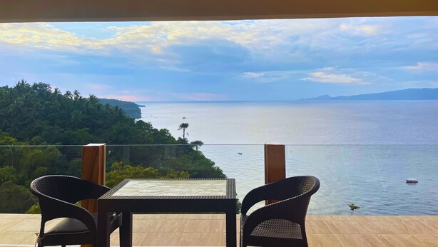 Un balcón con dos sillas de mimbre y una mesa, con vista a un mar tranquilo y una ladera boscosa.