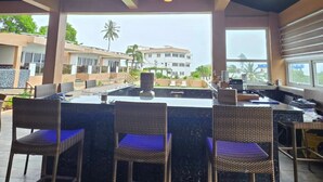 Zona de bar al aire libre con sillas de mimbre y una barra central, con vistas a una zona residencial con palmeras y casas.