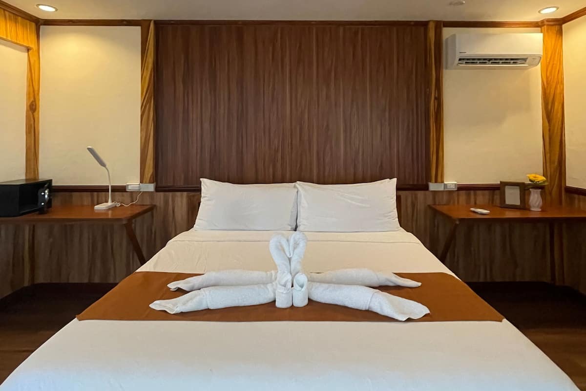 A hotel room with a large bed, wooden headboard, two bedside tables with lamps, and an air conditioning unit on the wall.