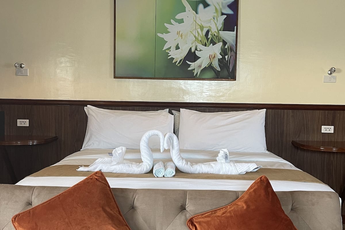 A hotel room with a large bed, two brown pillows, a wall-mounted painting of flowers, and a wooden headboard.