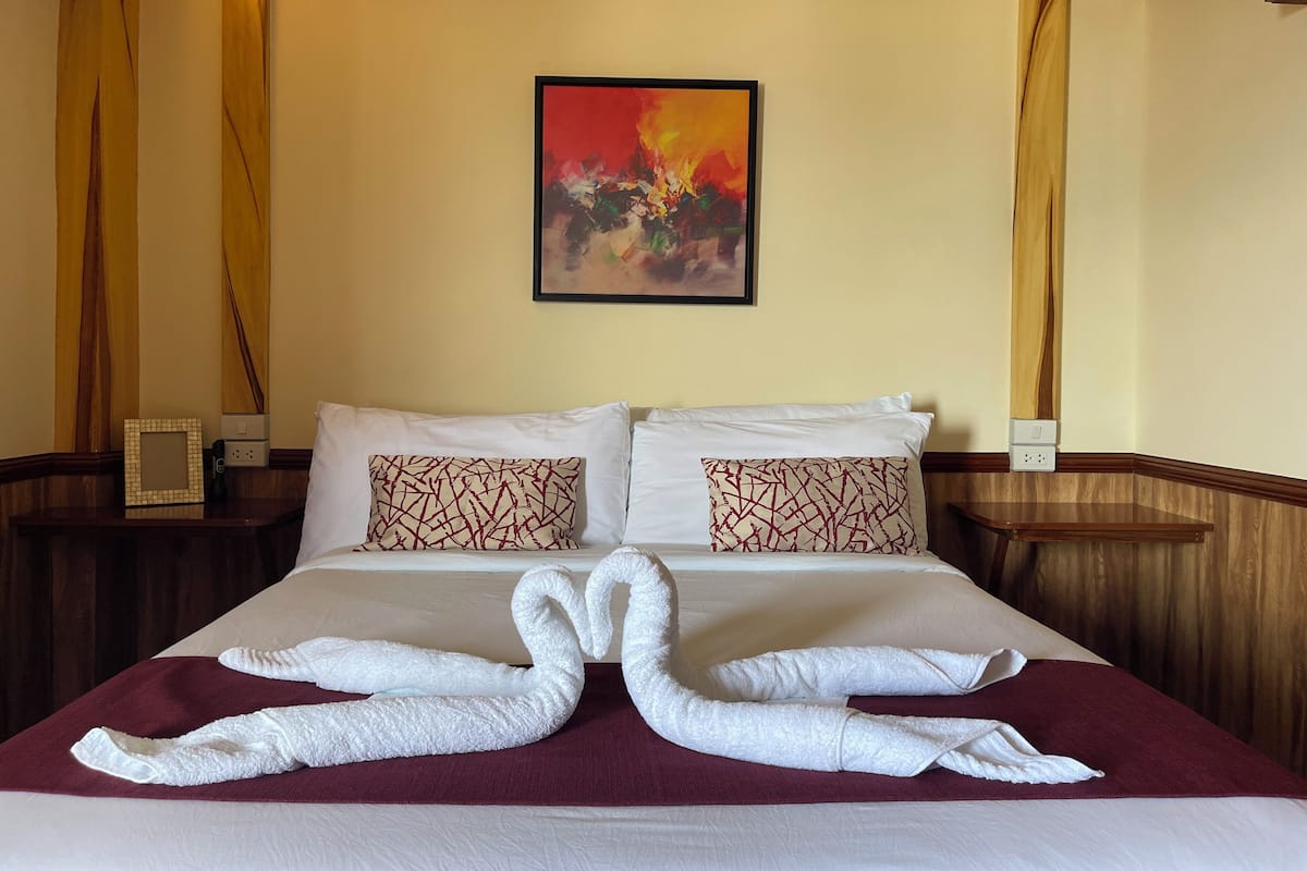 A hotel room with a bed, two pillows, a framed painting, and a swan-shaped towel arrangement.