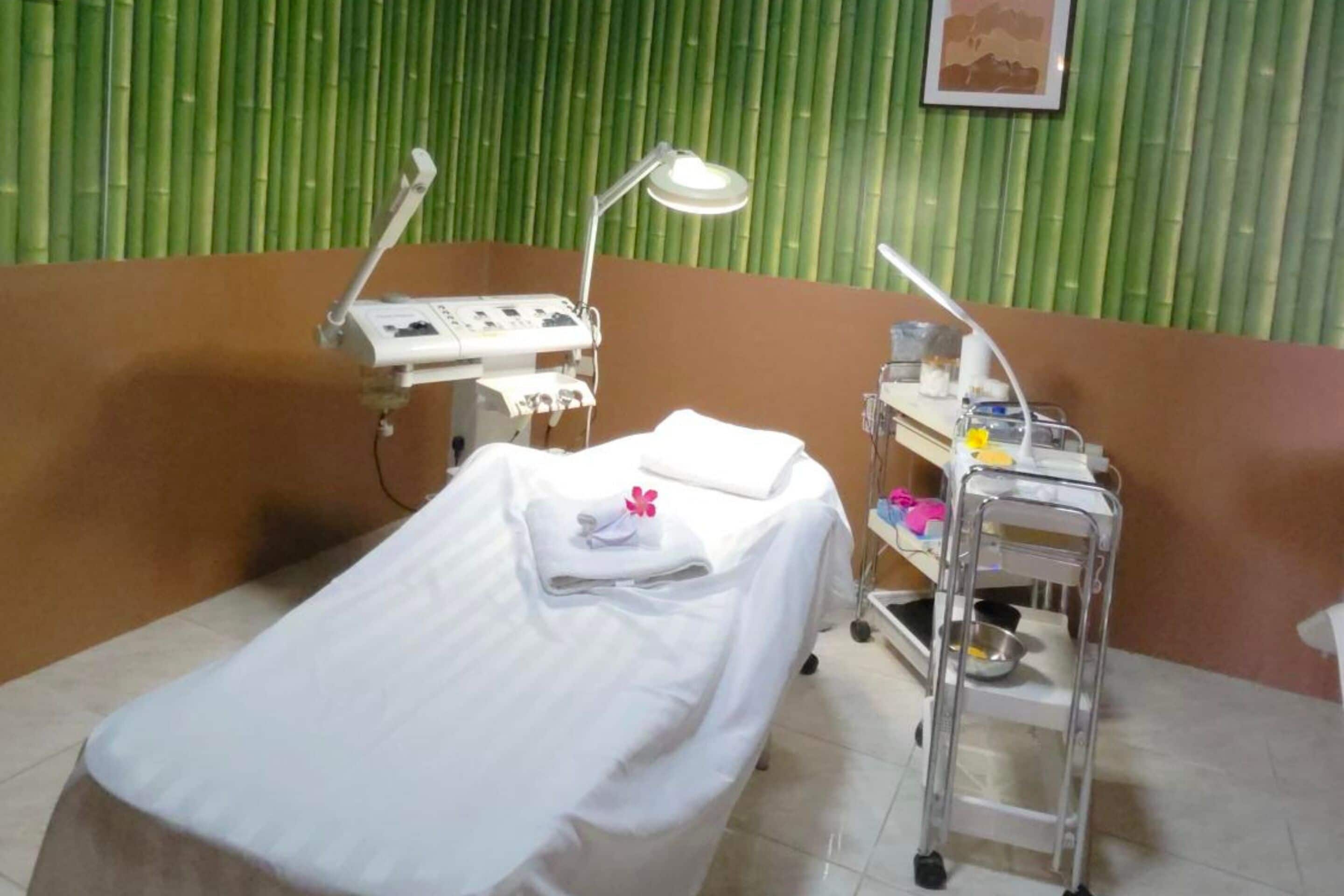 A medical examination room with a bed, two examination lamps, a medical cart, and a green bamboo wall.