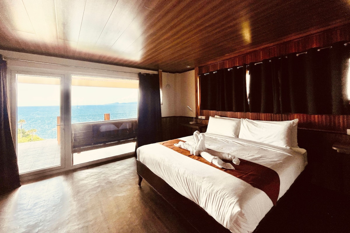 A bedroom with a large bed, a wooden ceiling, and a view of the sea through a sliding door.