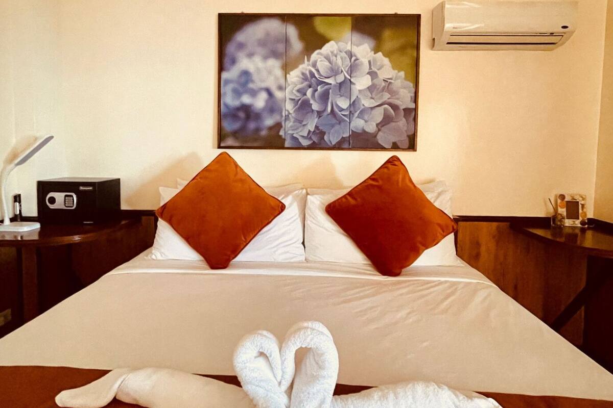 A hotel room with a neatly made bed, a painting of flowers on the wall, and an air conditioning unit.