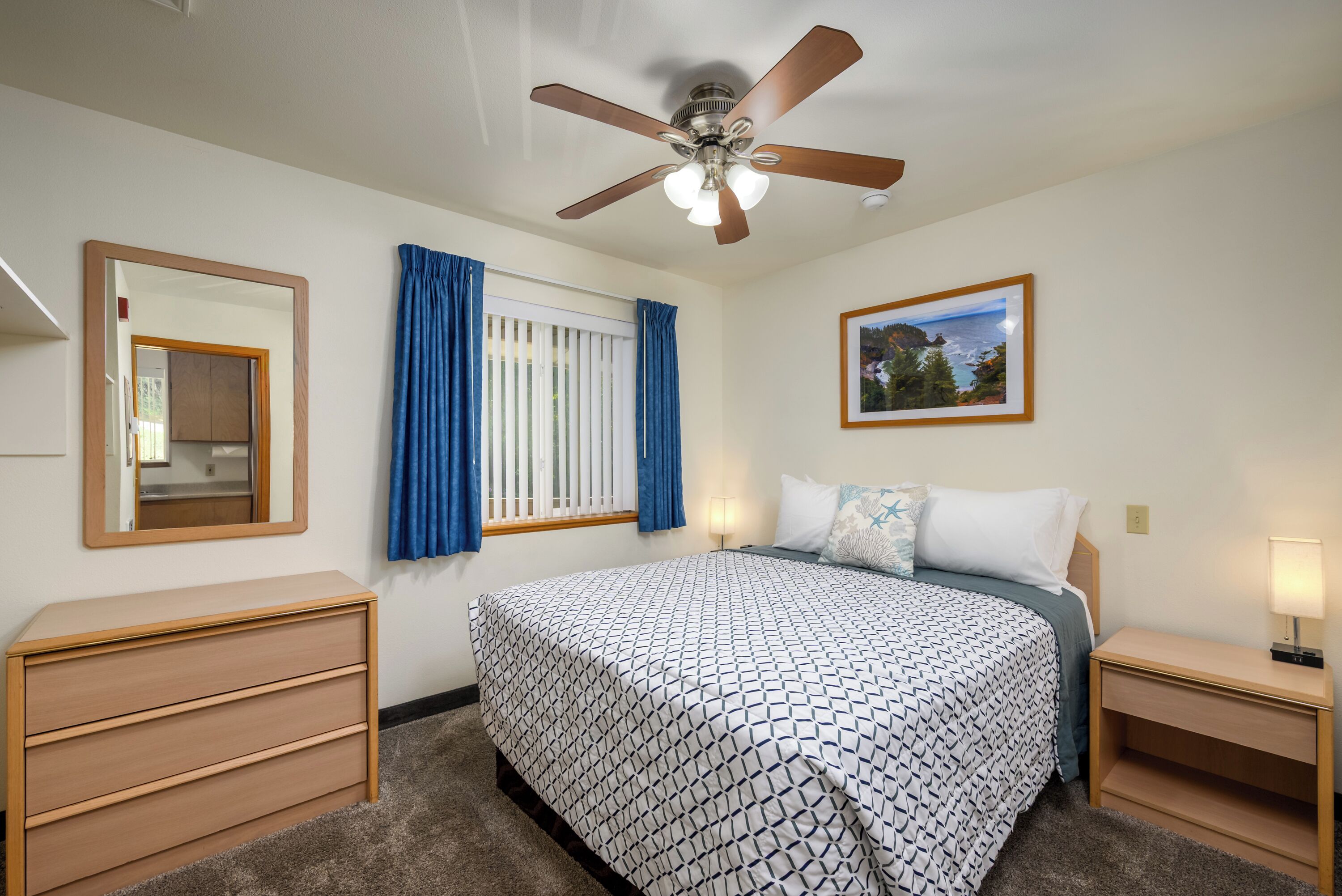 A neatly arranged bedroom with a bed, bedside tables, a ceiling fan, a window with curtains, a mirror, and a framed picture on the wall.