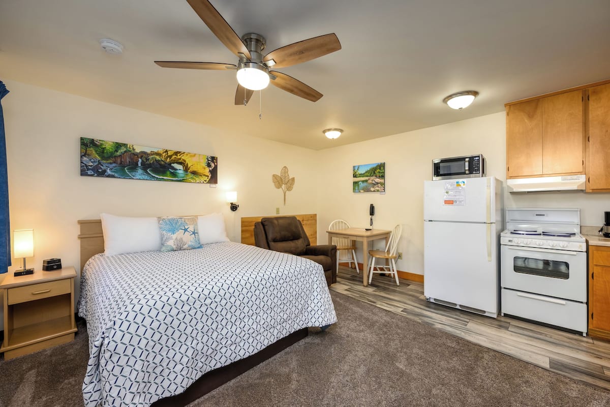 A hotel room with a bed, a desk, a chair, a microwave, a refrigerator, and a ceiling fan.