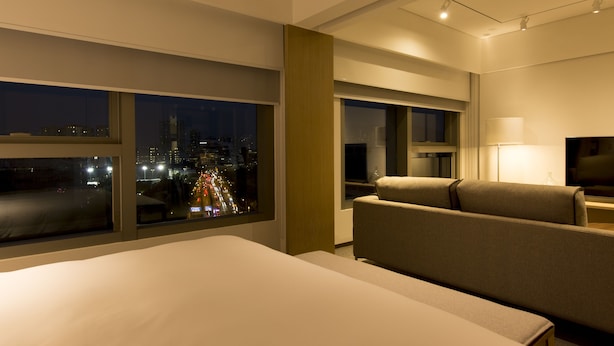 Habitación de hotel con una cama grande, un sofá, un televisor y una ventana con vista a la ciudad de noche.