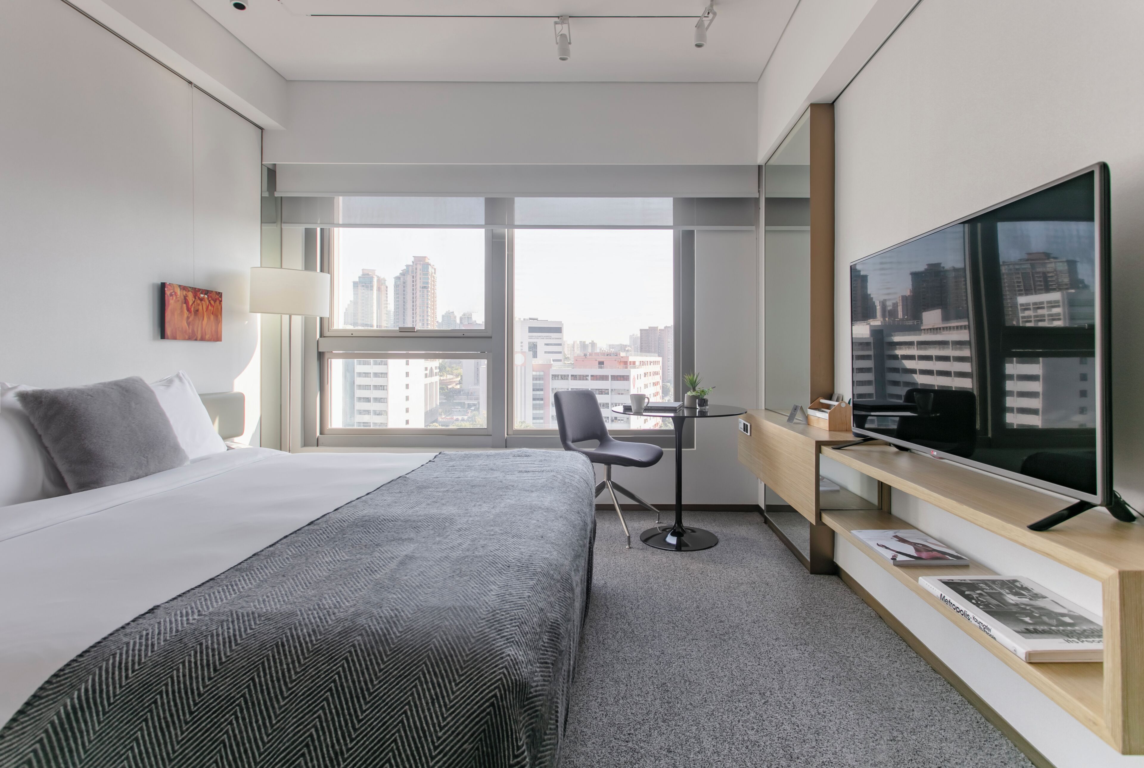 A hotel room with a bed, a desk, a chair, a TV, and a large window with a city view.