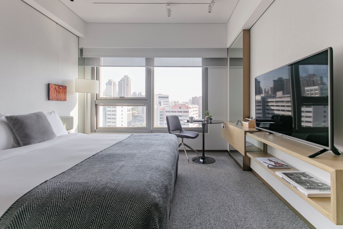 Una habitación de hotel moderna con una cama grande, un escritorio, un televisor y vistas a la ciudad a través de amplias ventanas.
