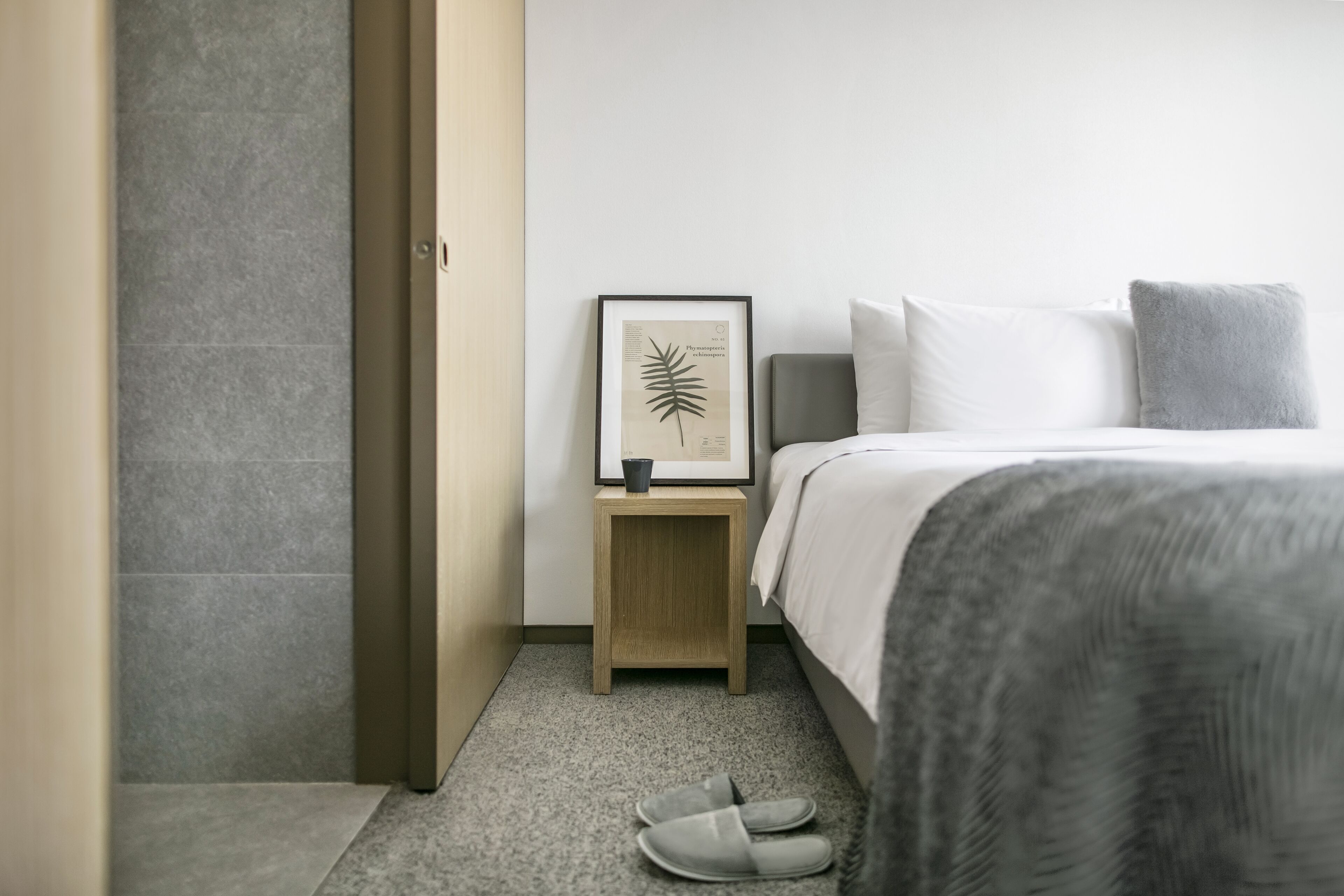 Una camera da letto ordinata con un letto, un comodino con una stampa botanica incorniciata e un paio di ciabatte sul pavimento.
