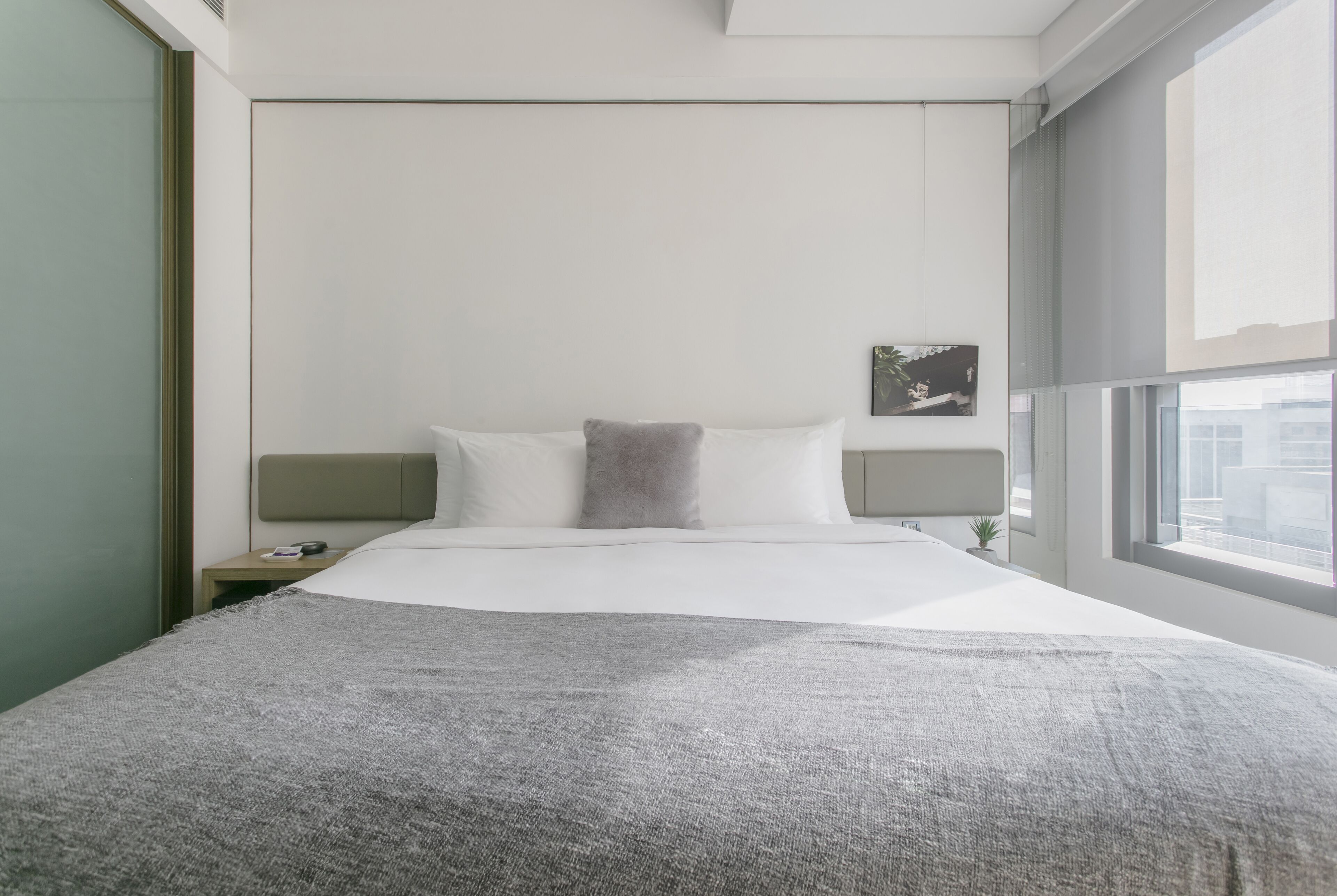 A modern hotel room with a large bed, a nightstand, a small plant, and a window with a view of buildings.