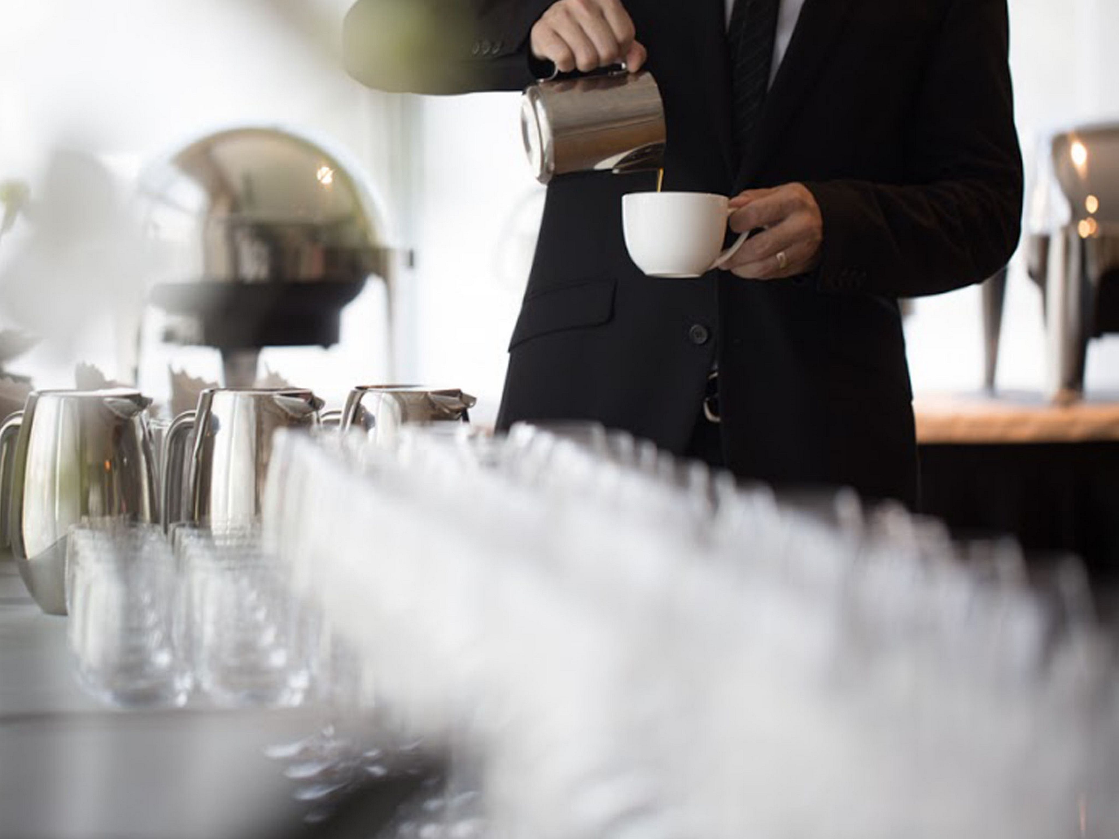 Une personne verse du café dans une tasse, sur une table où se trouvent une machine à café et d’autres tasses.