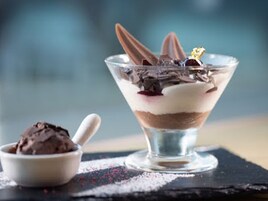A dessert in a glass bowl with layers of chocolate and cream, a spoon, and a small bowl of chocolate ice cream on a slate surface.