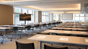 A conference room with rows of tables and chairs, large windows, a projector screen, and a whiteboard.