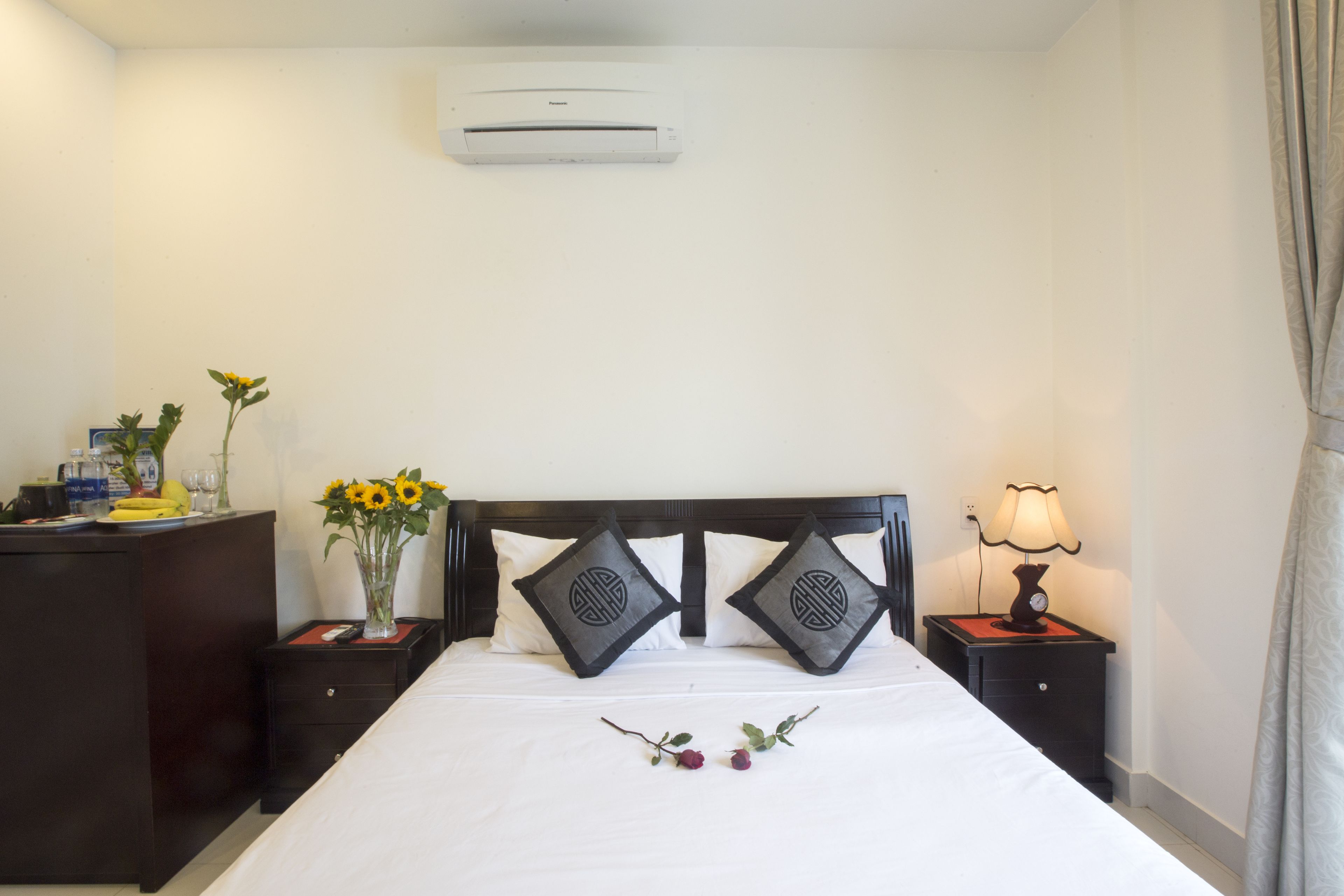 A hotel room with a bed, a desk, a nightstand, a lamp, a vase with flowers, and an air conditioner.