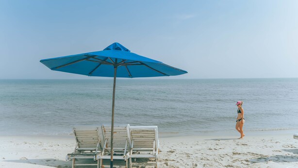 A beach with a blue umbrella, two white lounge chairs, and a person in a bikini walking along the shore.