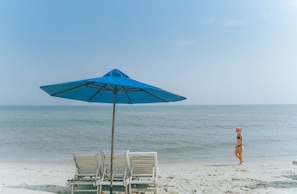 Een strand met een blauwe parasol, twee witte ligstoelen en een persoon in een bikini die langs de kust loopt.