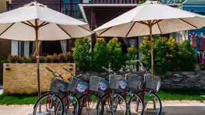 A row of bicycles with baskets, each with a delivery box attached, parked under umbrellas in an outdoor setting.