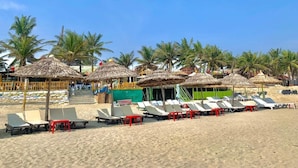 A beach with rows of sun loungers and thatched umbrellas, palm trees, and a building with a green awning.