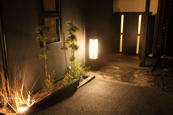 Vista nocturna de un sendero estrecho que conduce a la entrada de un edificio, bordeado por macetas y una lámpara de pared iluminada.