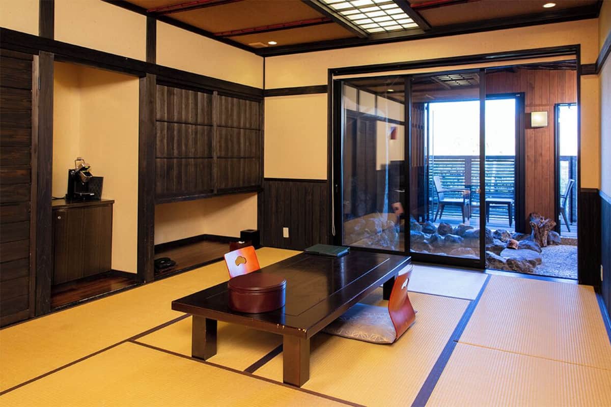 Ein traditioneller japanischer Raum mit Tatami-Böden, einem niedrigen Tisch und Schiebetüren, die zu einem Außenbereich mit Blick auf Felsen und eine Holzterrasse führen.
