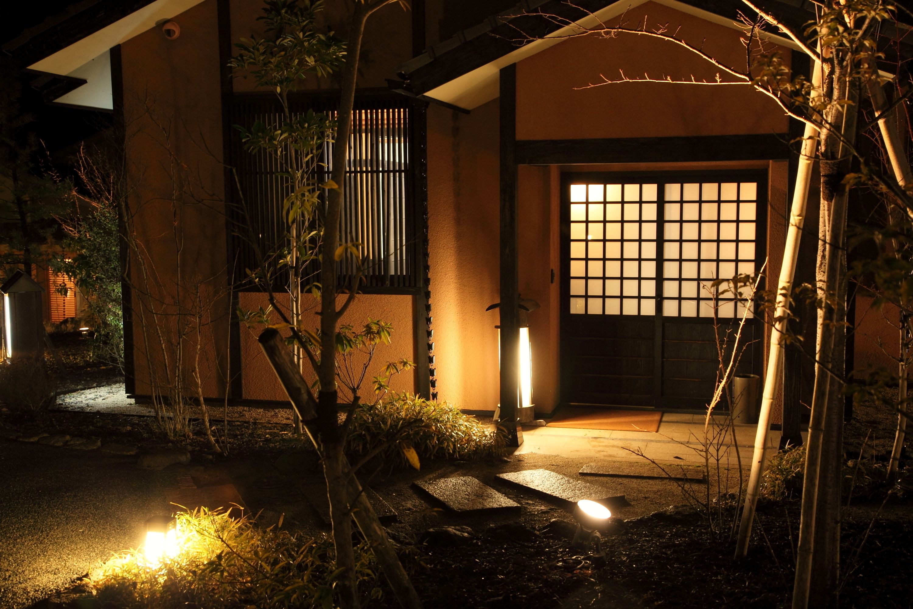 A traditional Japanese building at night with a wooden gate, illuminated by ground lights, surrounded by bamboo and shrubs.