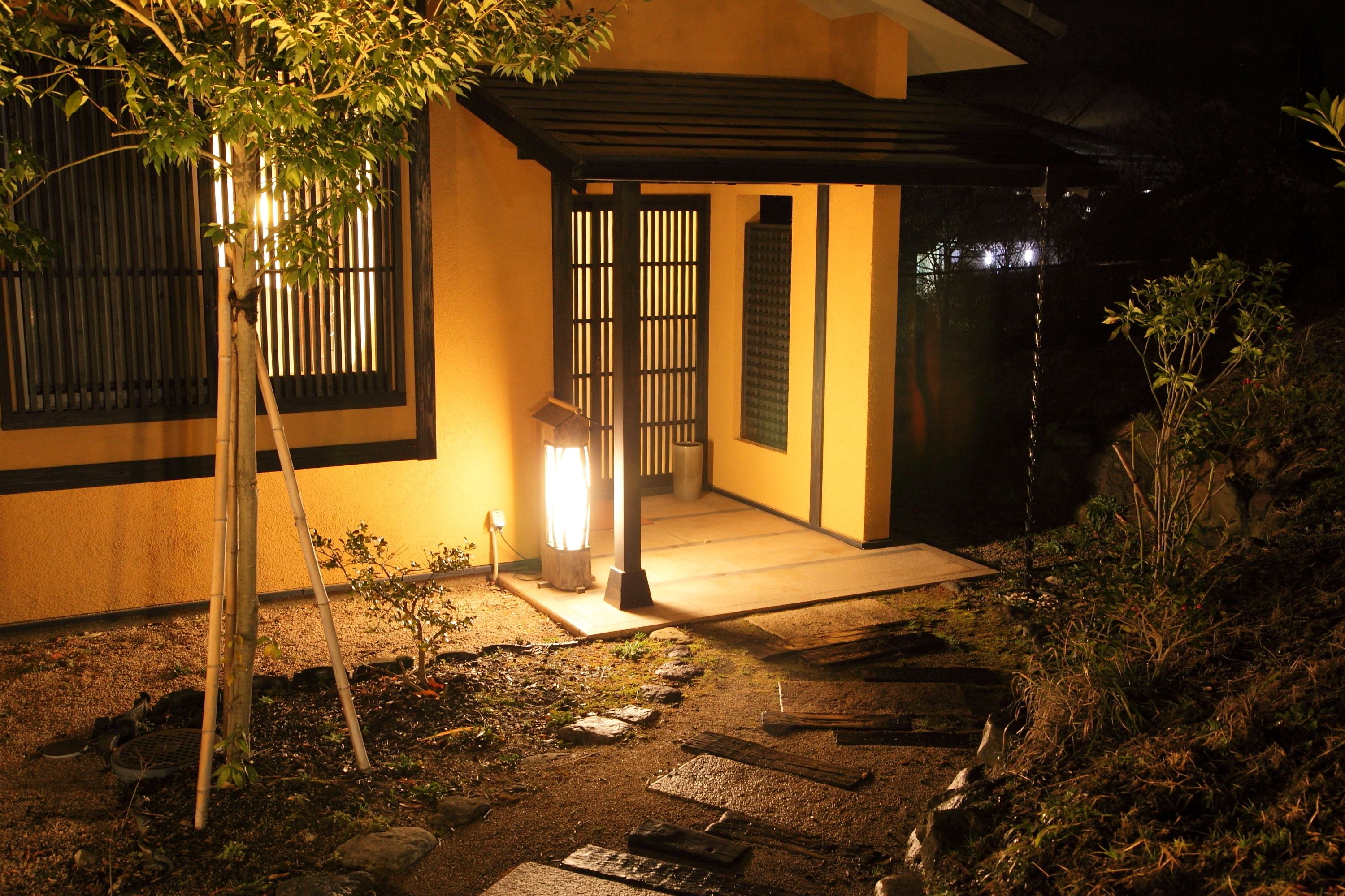 A traditional Japanese-style entrance with a wooden gate, a stone pathway, and a small tree.