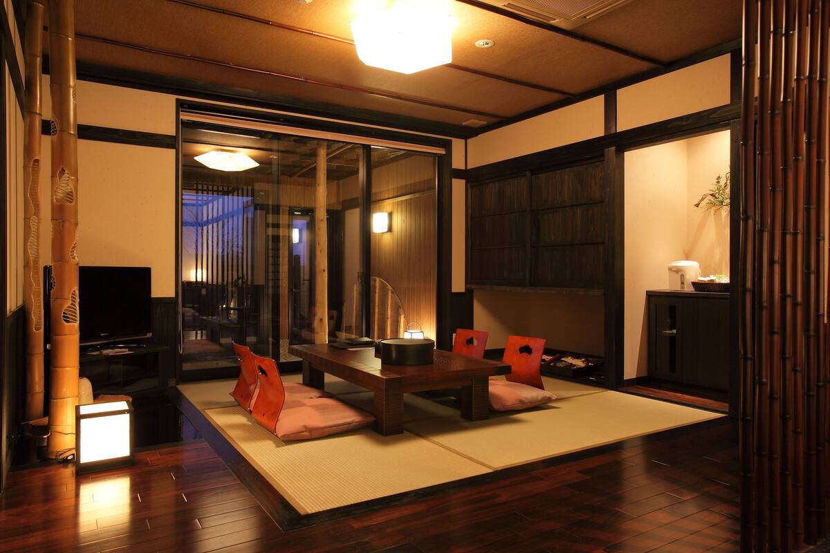 Ein traditioneller japanischer Raum mit Tatami-Bodenbelag, einem niedrigen Tisch mit Kissen, einem Fernseher und verschiebbaren Türen.