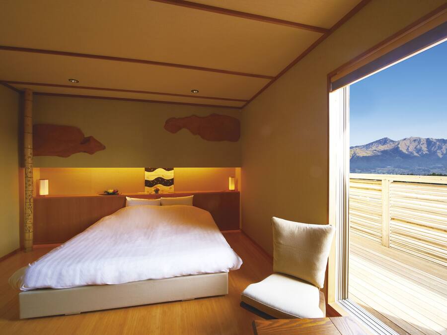 Habitación de hotel con una cama grande, una silla, una mesita de noche y vistas a montañas a través de una ventana.
