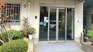 A building entrance with a glass door, a small garden area with potted plants, and a blue mat at the entrance. - HOTEL LiVEMAX BUDGET Kawasaki Daishi (Kawasaki)